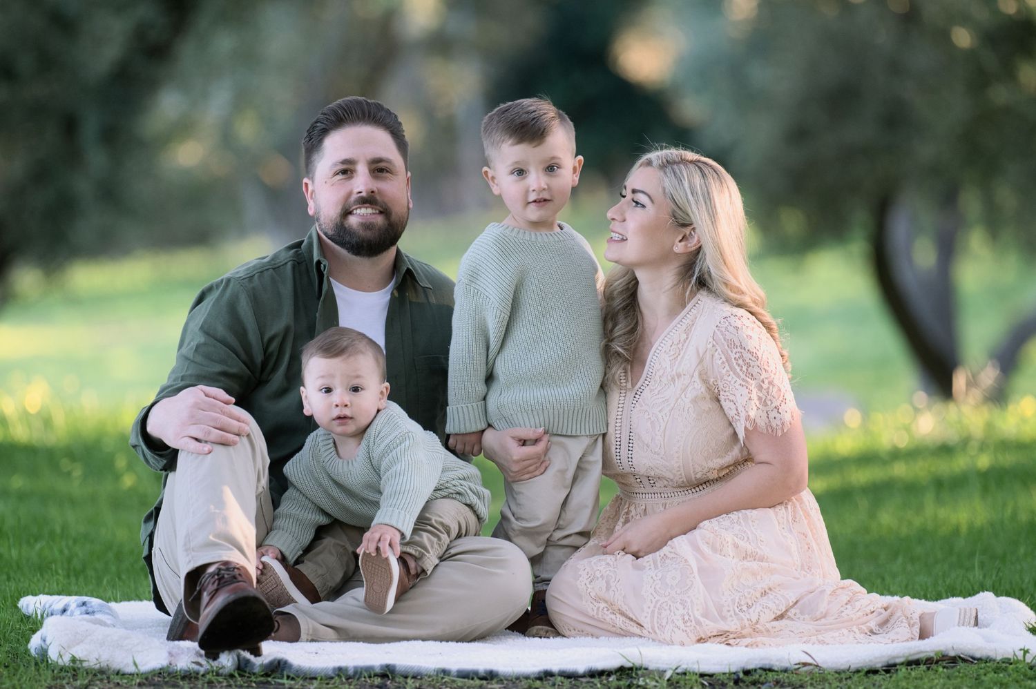 A happy family of four sits on a blanket in a sunny green park with olive trees.