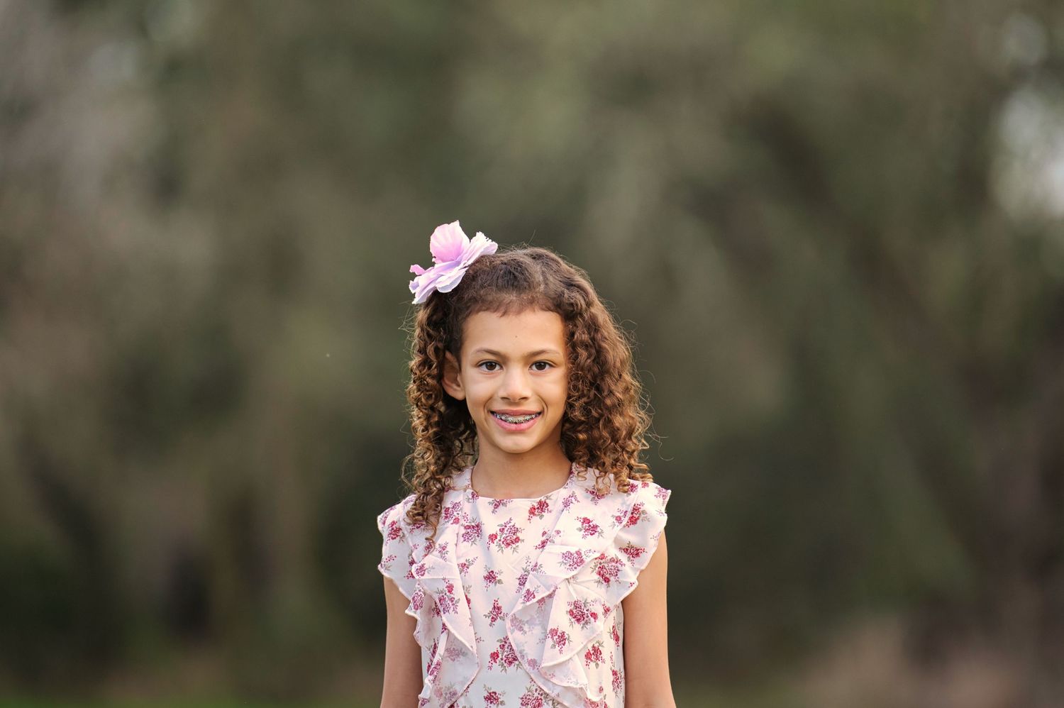 A young girl with curly hair and a pink bow smiles warmly in an outdoor portrait session.