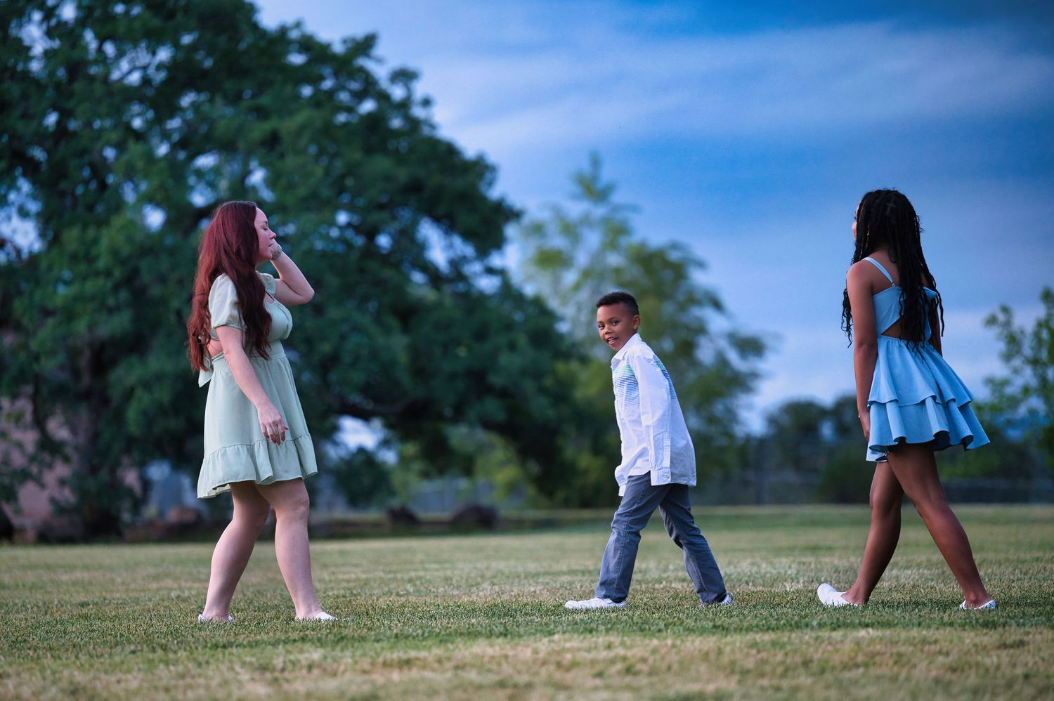 Two women in pastel dresses with a young boy walking playfully on a green lawn at dusk.
