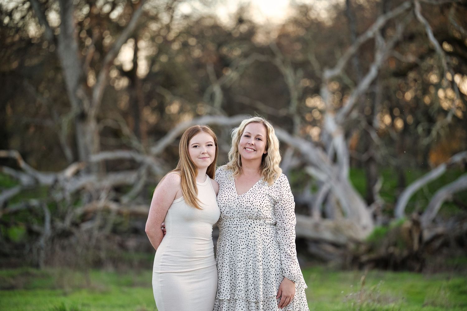 Two women smile together outdoors near fallen trees in a sunny field, one in white dress, one in floral.