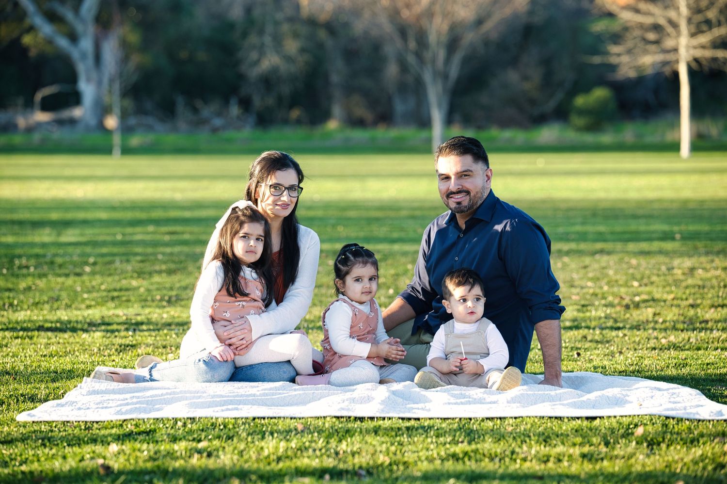 A happy family of five poses together on a white picnic blanket in a sunny green park.