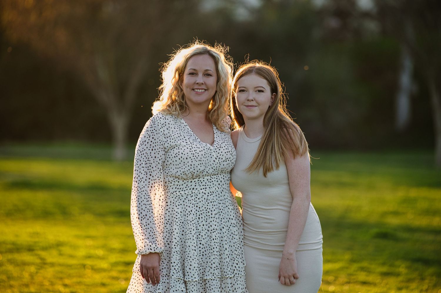 A mother and daughter pose together smiling in a sunlit green field wearing elegant dresses.