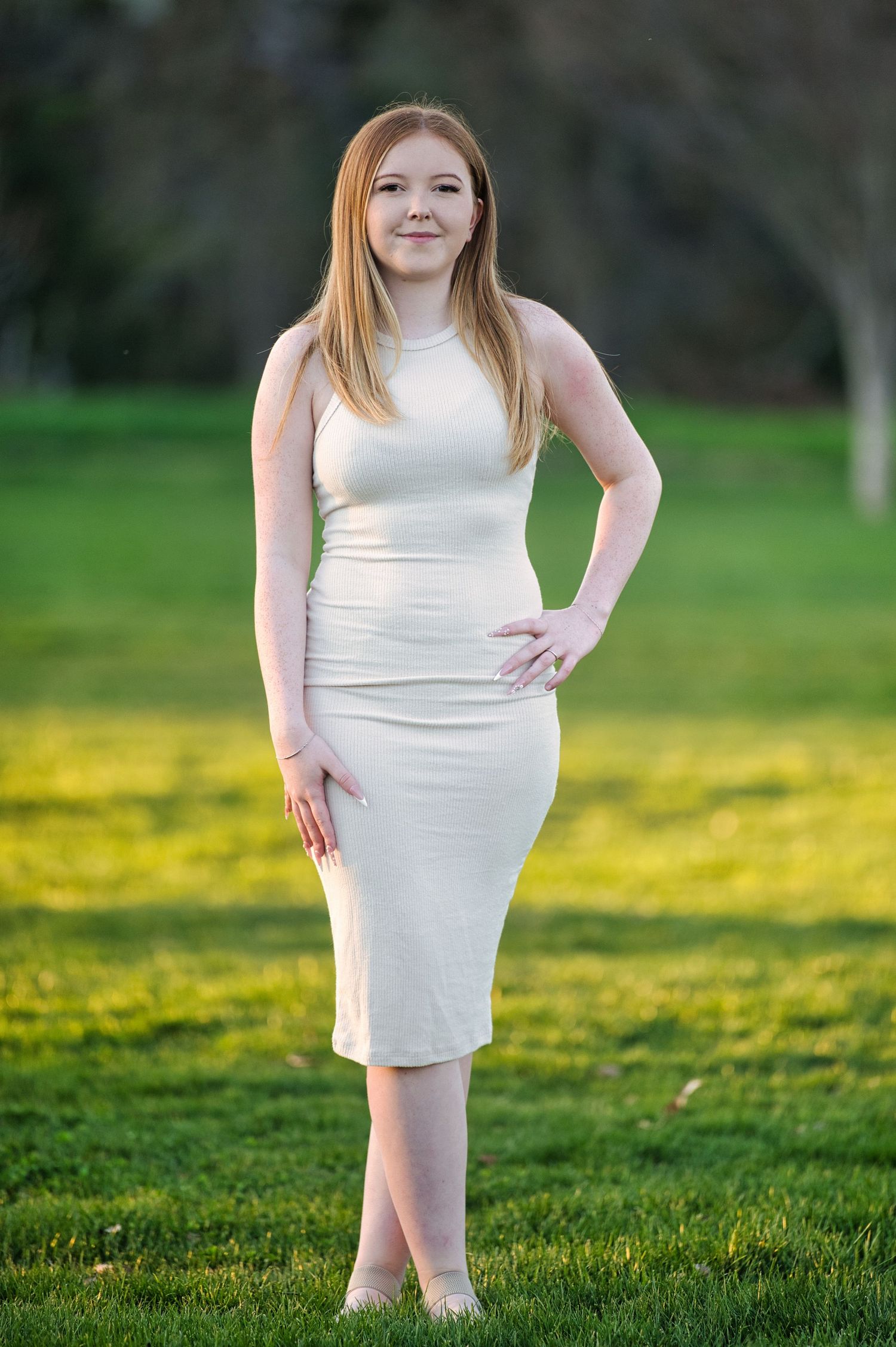 A young woman in a fitted cream dress poses confidently on a green lawn at sunset.