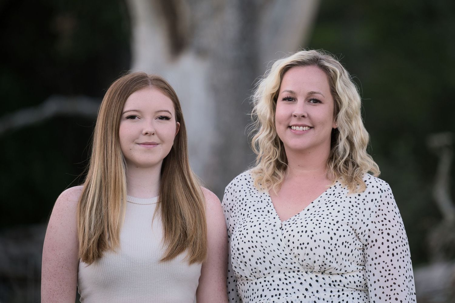 Two smiling women stand together outdoors in soft evening light near trees.