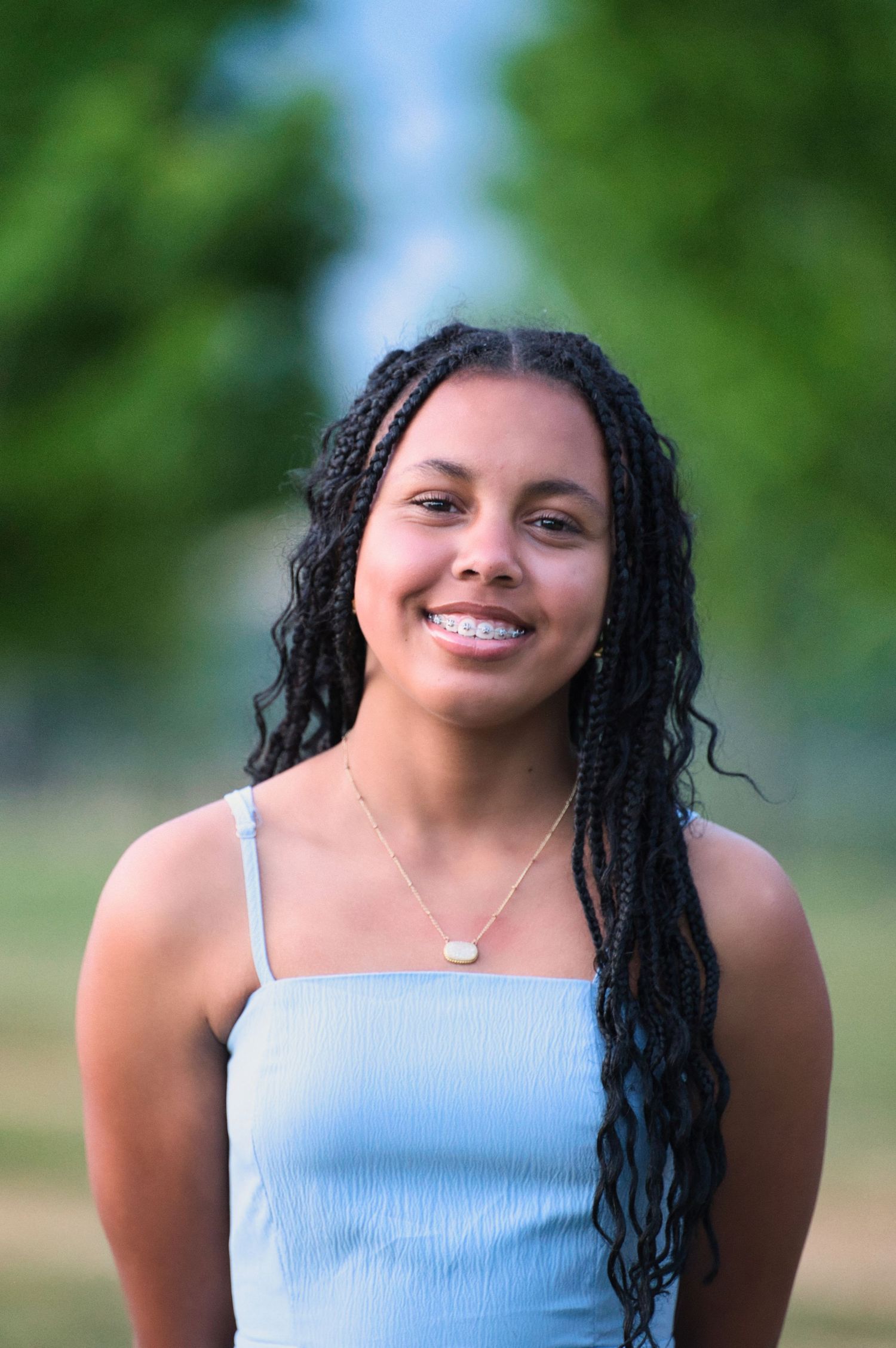 Smiling young woman with braided hair wearing a light blue top and gold necklace outdoors.