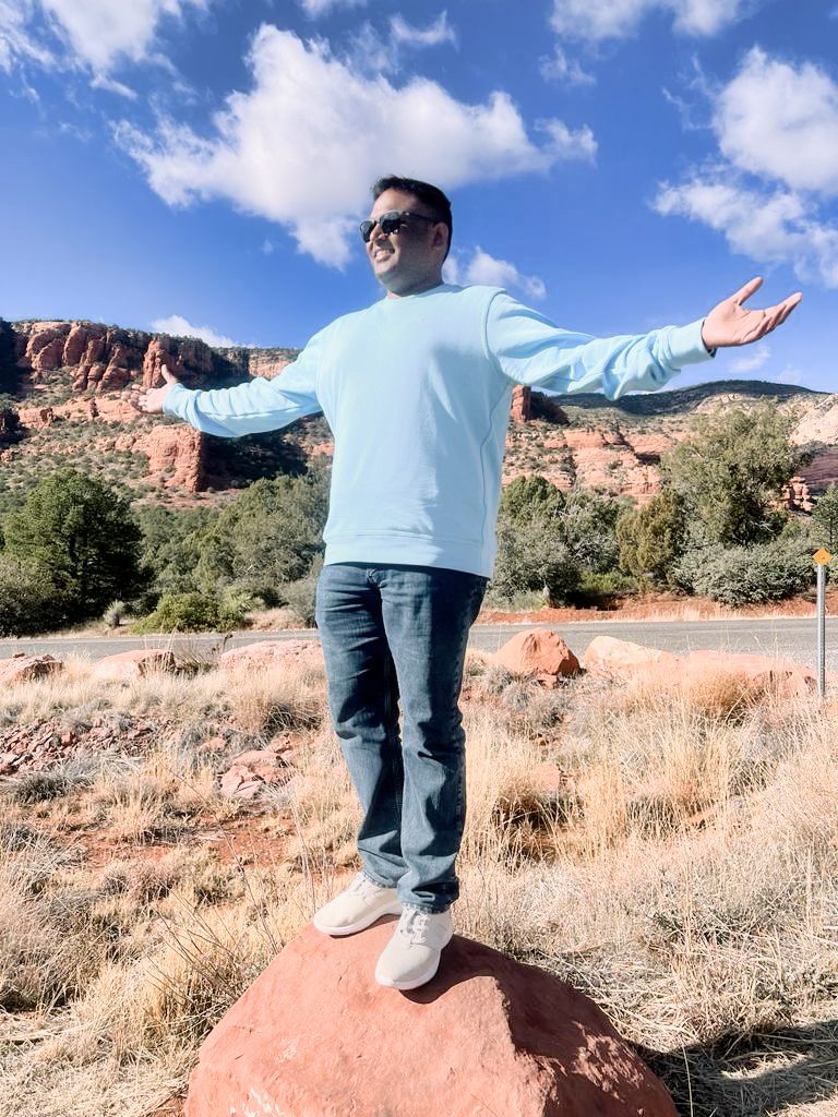 A man in a light blue sweater stands on a red rock with arms outstretched in Sedona, Arizona.
