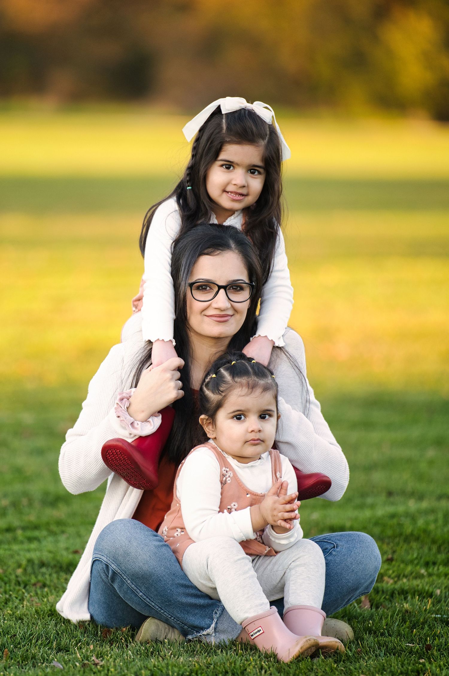 Mother with glasses sitting on grass with two young daughters in matching pink dresses.