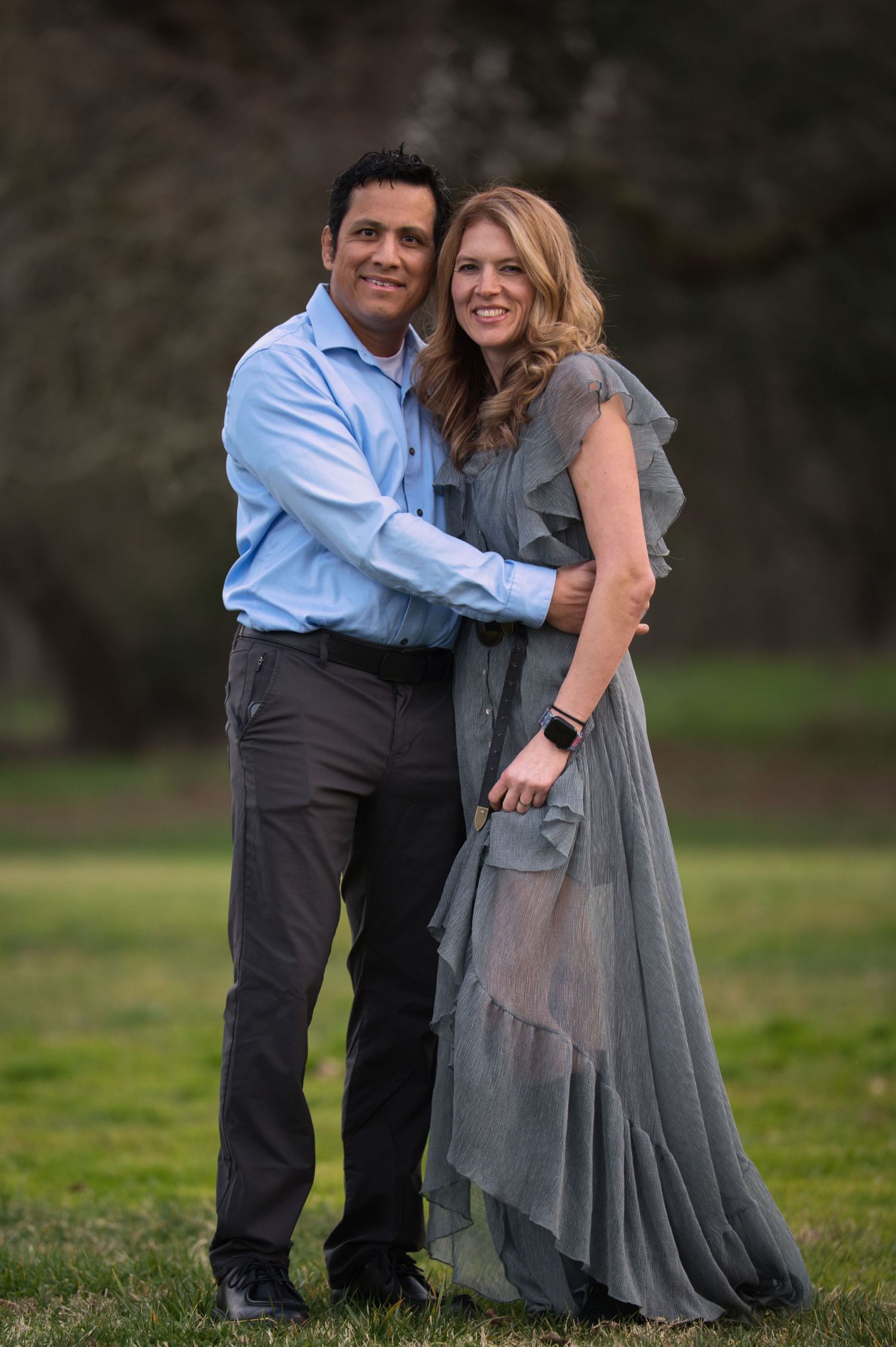 Smiling couple posing together on a green lawn, man in blue shirt embracing woman in grey ruffle dress.