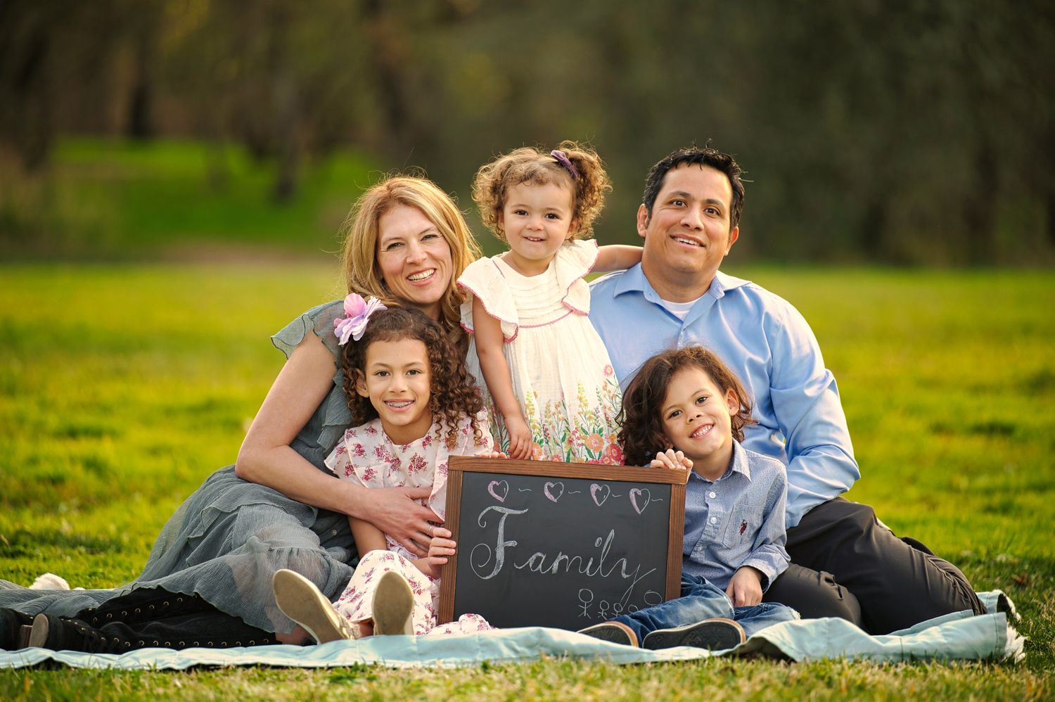 A cheerful family of five poses on grass in a park with a child holding a chalkboard Family sign.