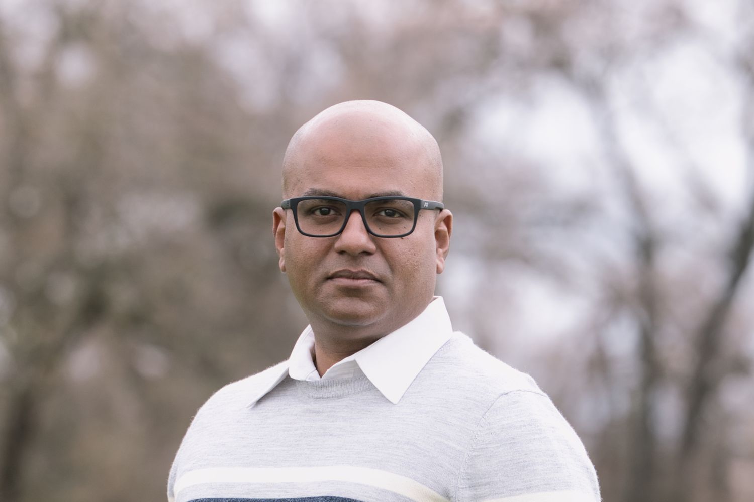 Close-up portrait of a bald man wearing glasses and a grey sweater outdoors in a park setting.
