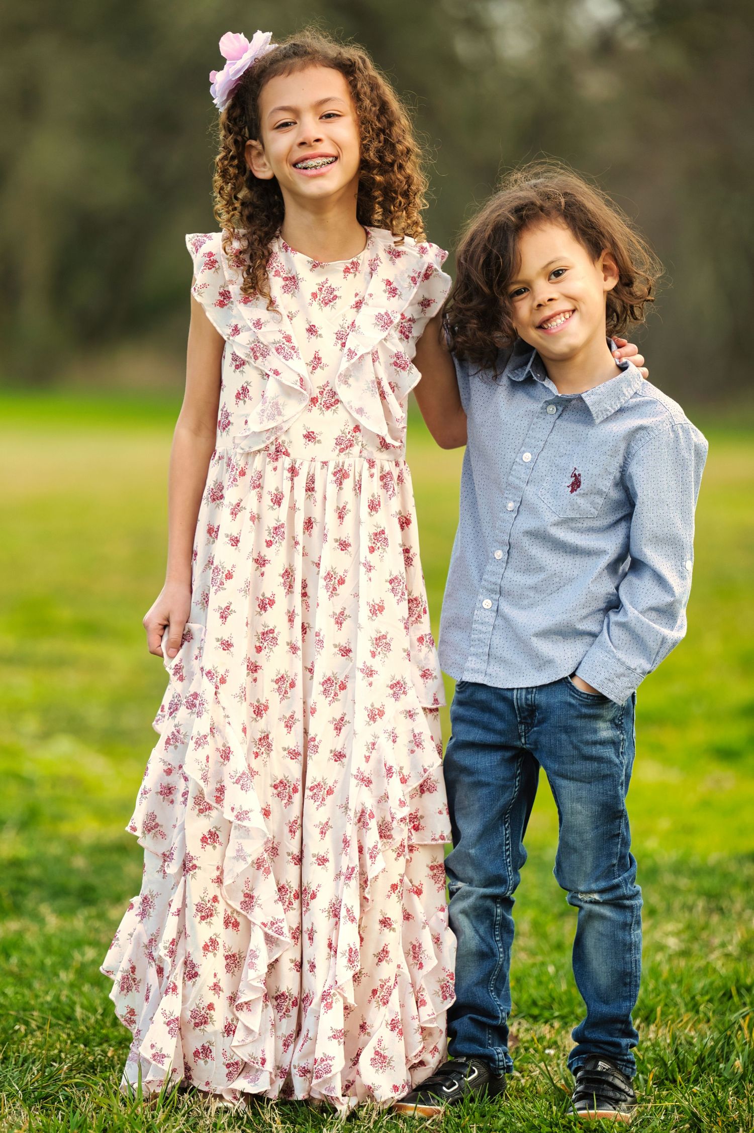 Smiling siblings in a park, girl wearing a floral ruffle dress with pink bow, boy in a blue button-up shirt.