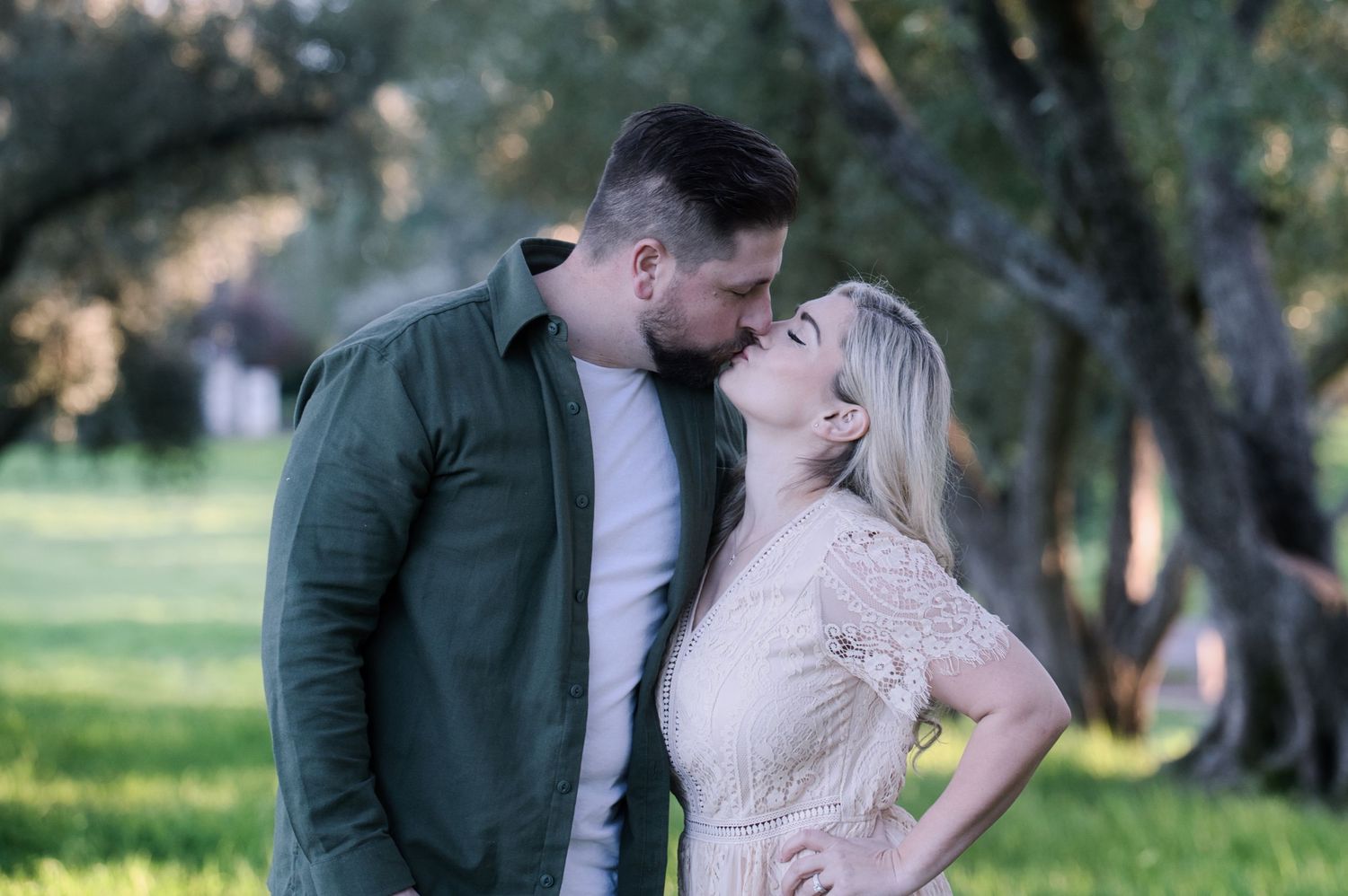 A couple shares a romantic kiss outdoors surrounded by lush green trees in soft natural light.