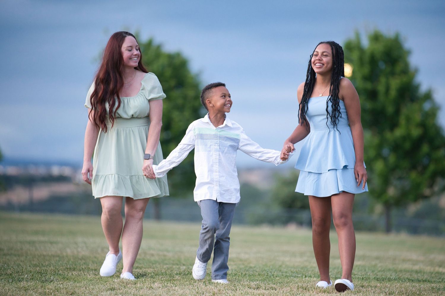 Two women in pastel dresses holding hands with a smiling young boy walking in a green park.