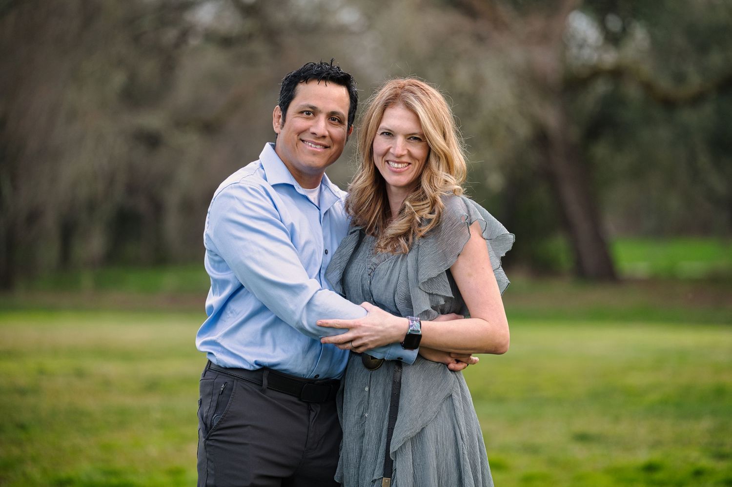 Smiling couple embracing outdoors in a green park with trees in background.