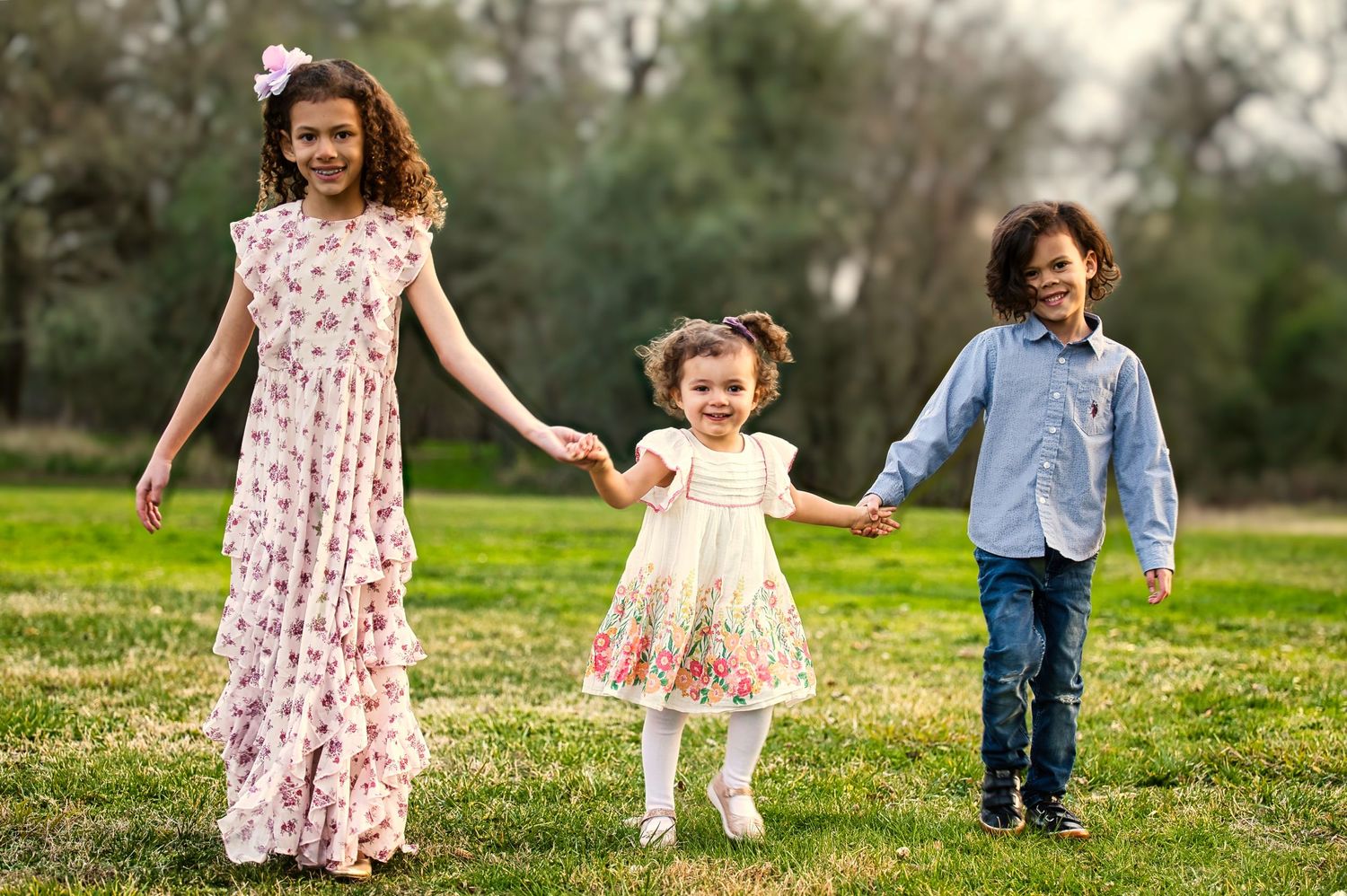 Three curly-haired siblings holding hands and walking on grass, girls in floral dresses, boy in blue shirt.