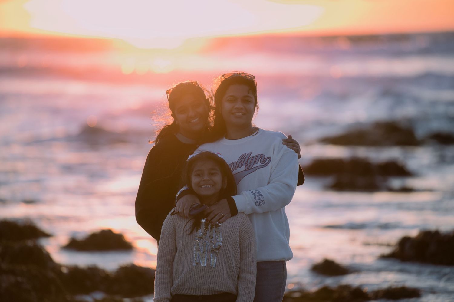 Three people embrace and smile together on a rocky beach during a vibrant golden sunset.