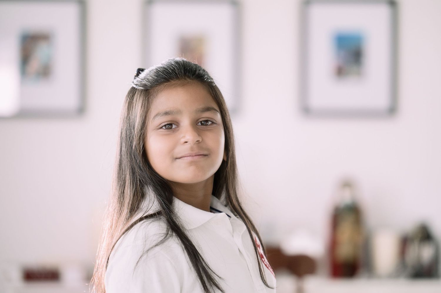 A young girl with long dark hair and a white bow smiles warmly indoors.