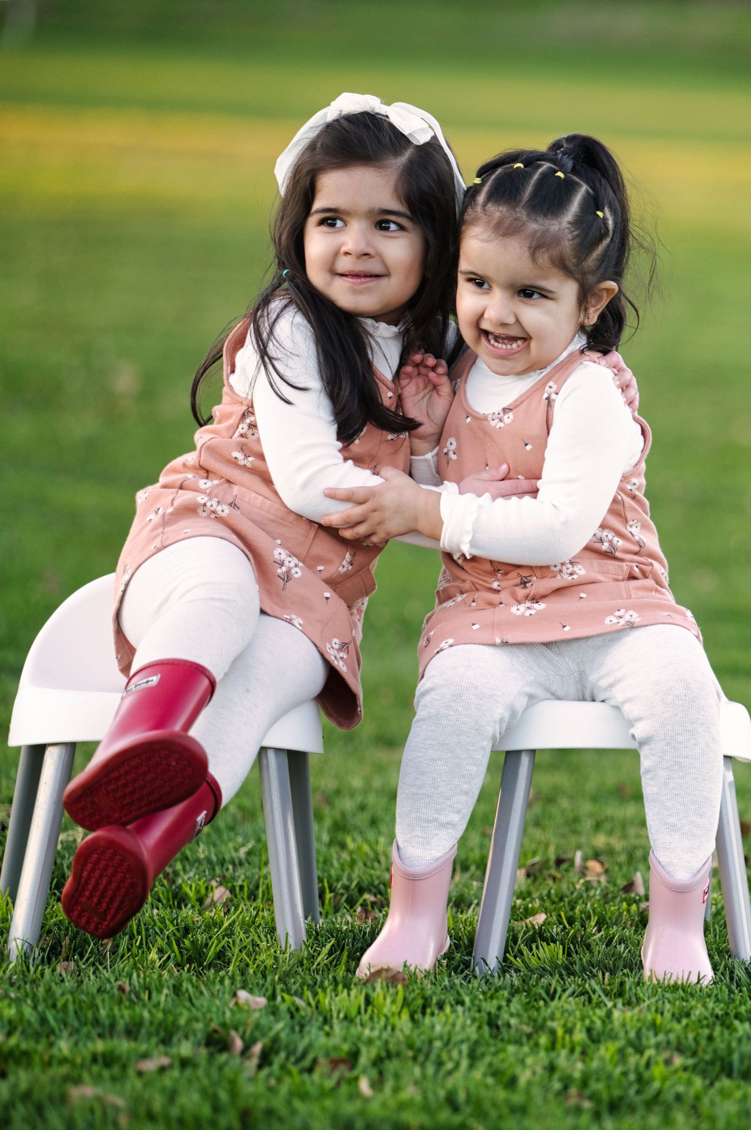 Two little sisters in matching pink dresses hugging on white chairs in green park.