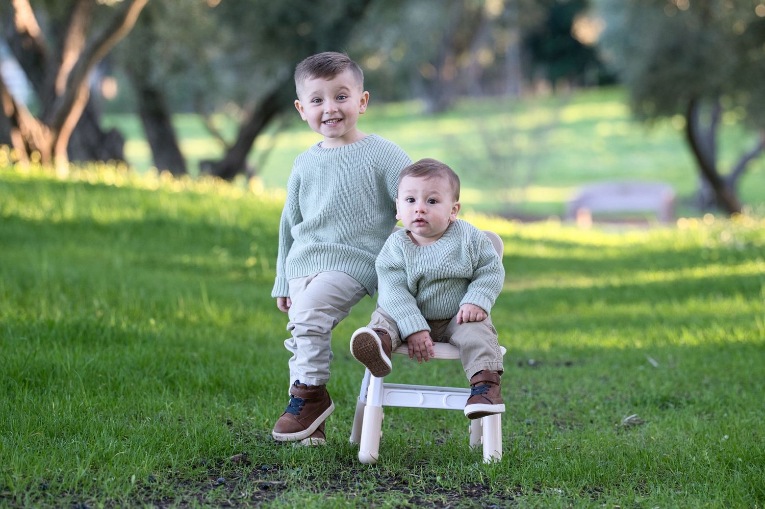 Two young brothers in matching sage green sweaters posing outdoors, older boy standing beside baby on small white chair.