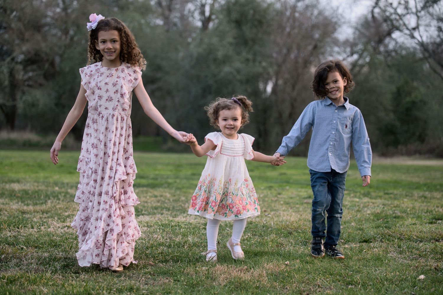 Three happy children in dressy outfits hold hands and walk together across a grassy park.