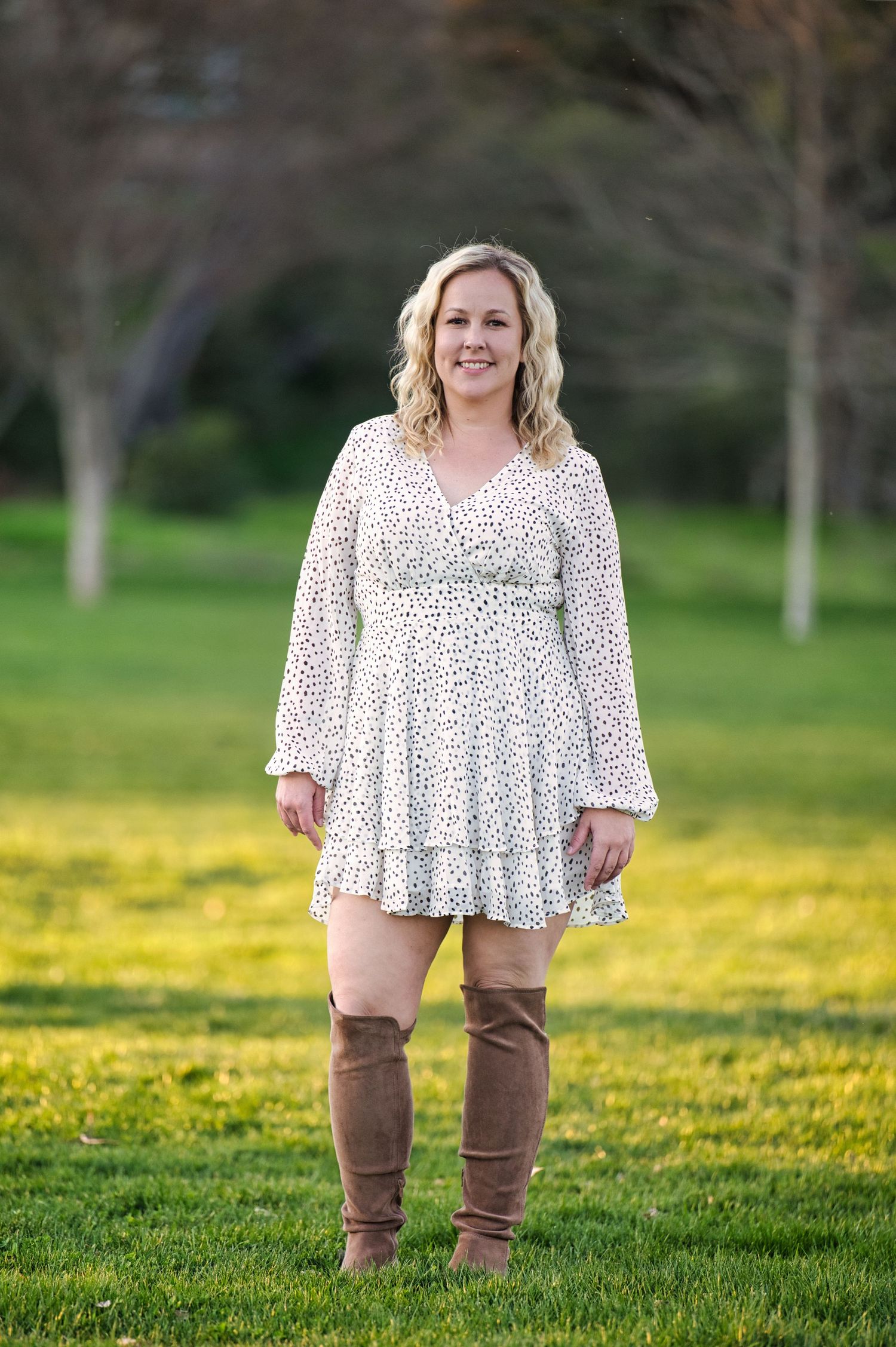 A smiling blonde woman in a white floral dress and brown knee-high boots stands in a park.