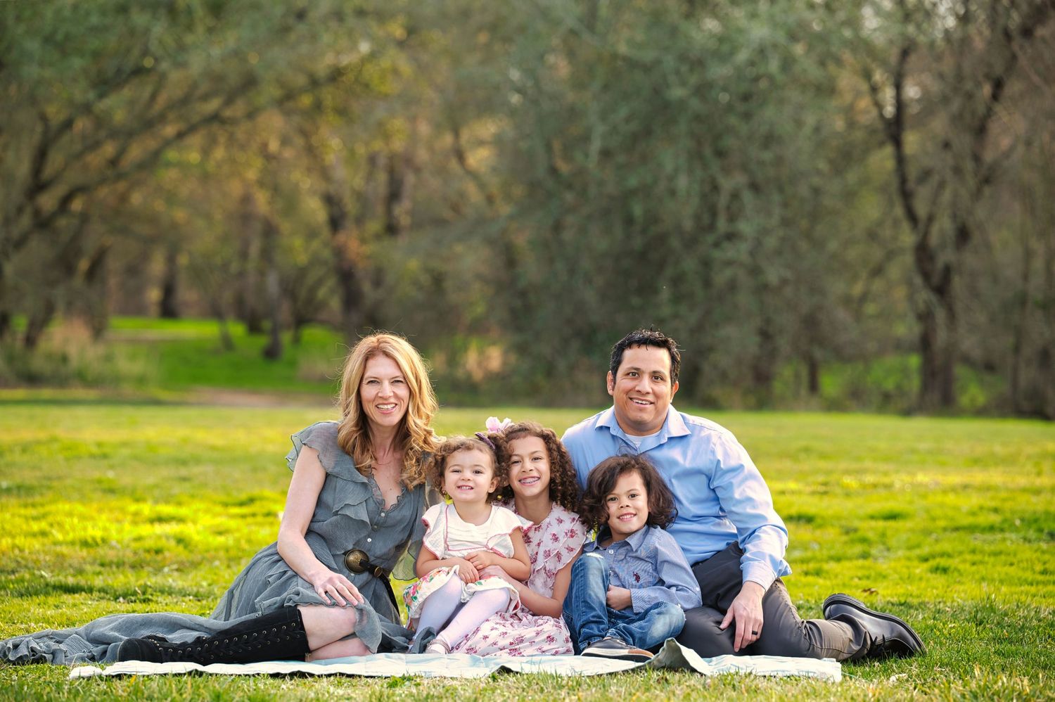 A family of five relaxes together on a white blanket in a lush green park surrounded by trees.