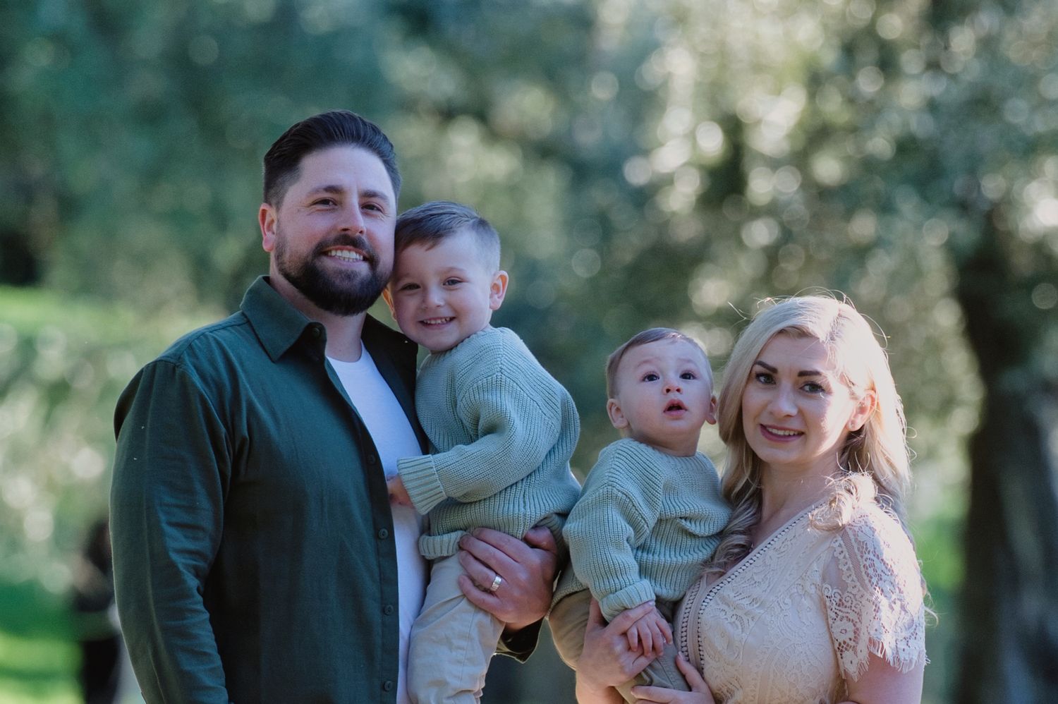 Happy family of four posing outdoors among olive trees, parents holding two young boys in matching green sweaters.