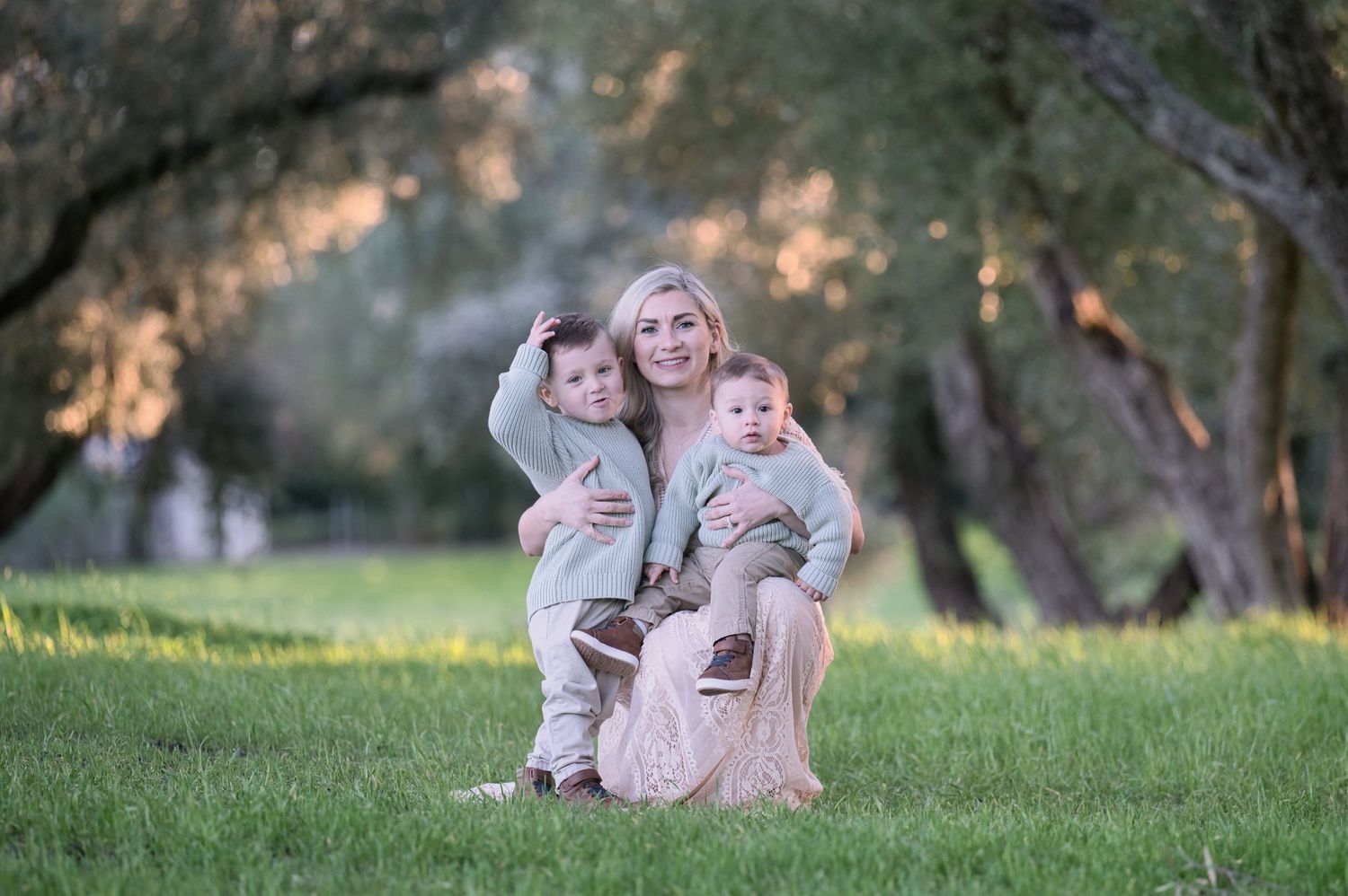 Blonde mother sitting on grass hugging two young boys in matching sage green sweaters during outdoor family photo session.