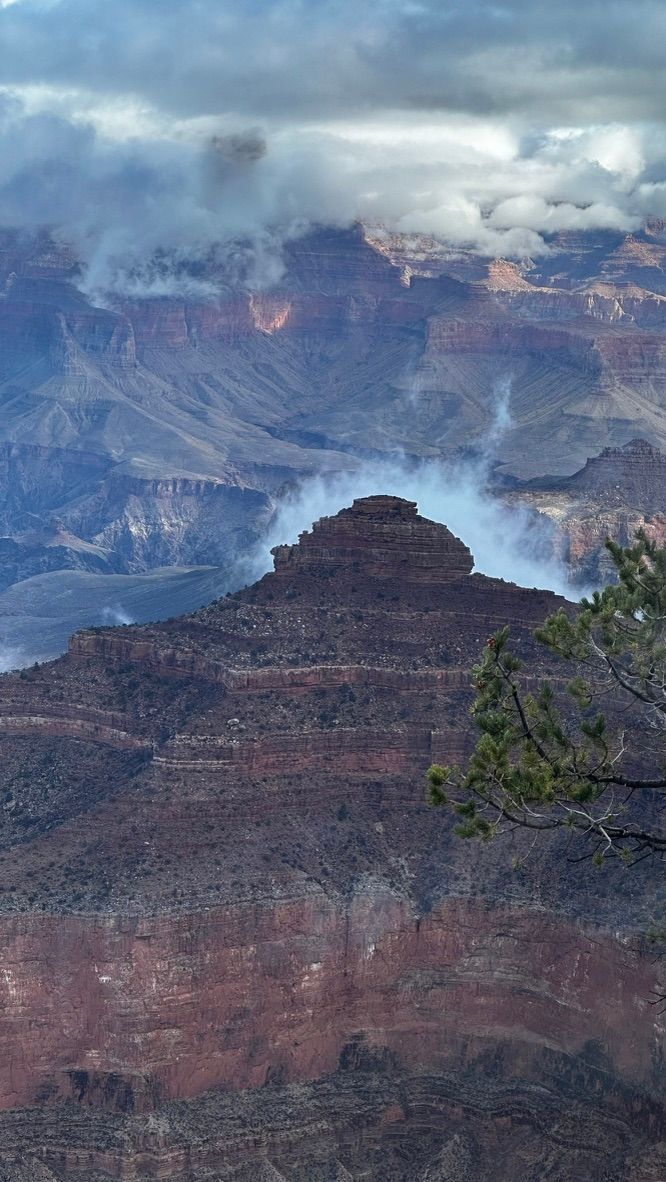 Dramatic clouds and smoke drift over the layered red rock formations of the Grand Canyon.