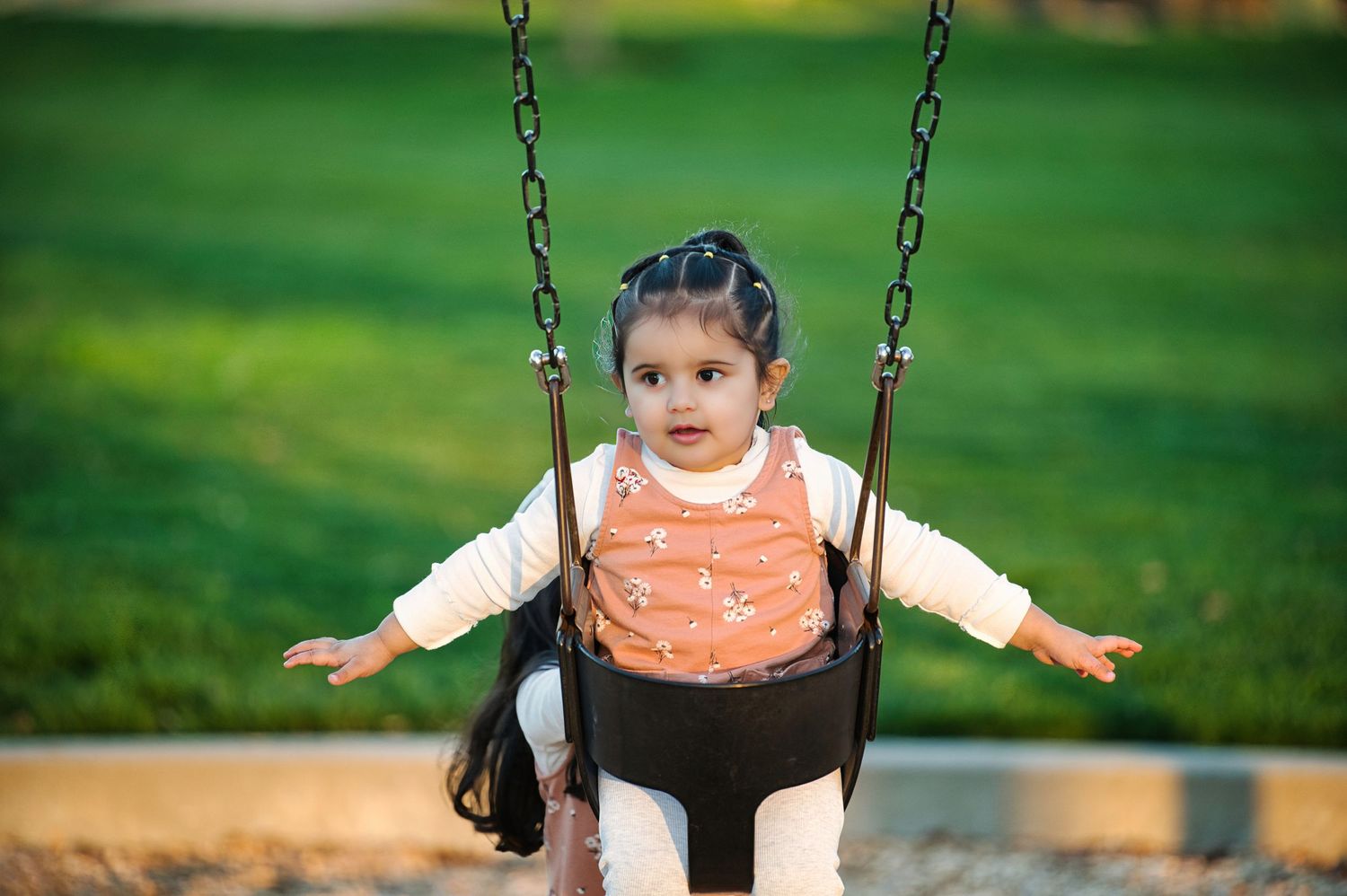 Toddler girl in pink vest sitting in playground swing with green grass background.