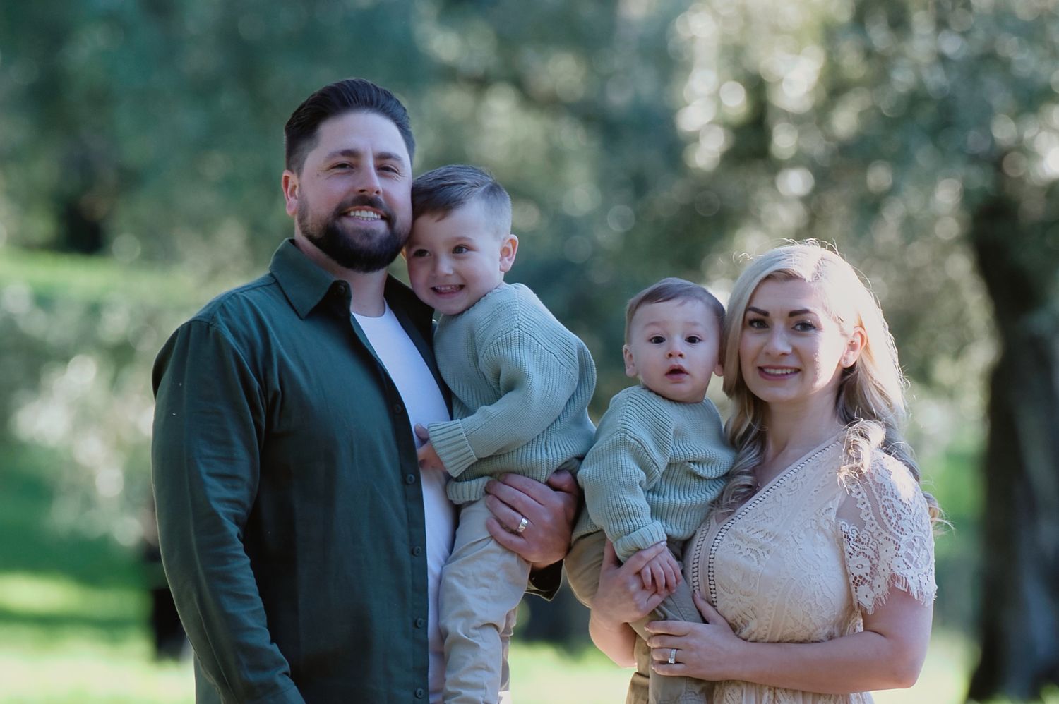 Family of four smiling outdoors among olive trees, parents holding two young boys in coordinating neutral outfits.