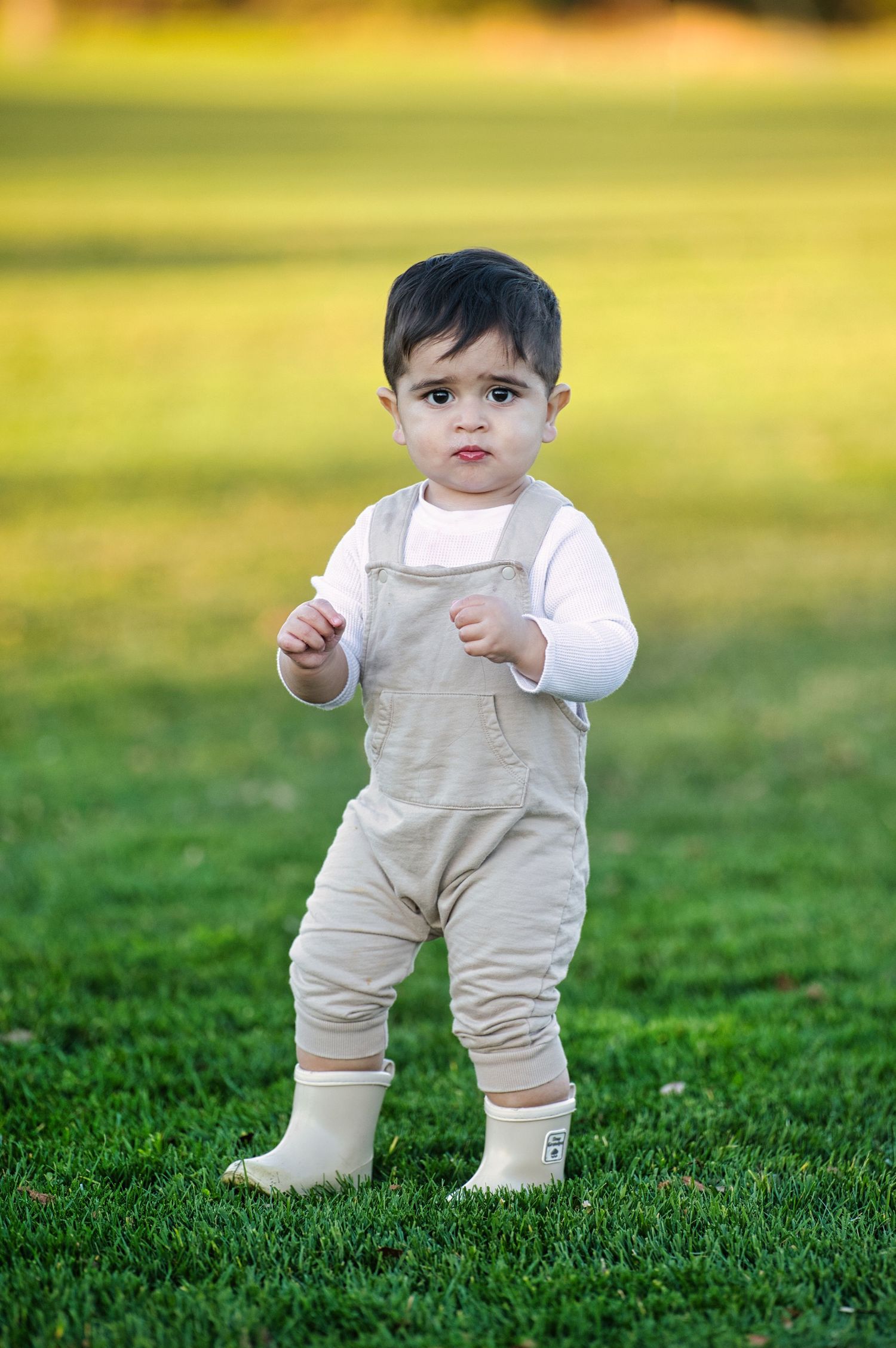 An adorable toddler in beige overalls and white long sleeves stands on a lush green lawn.