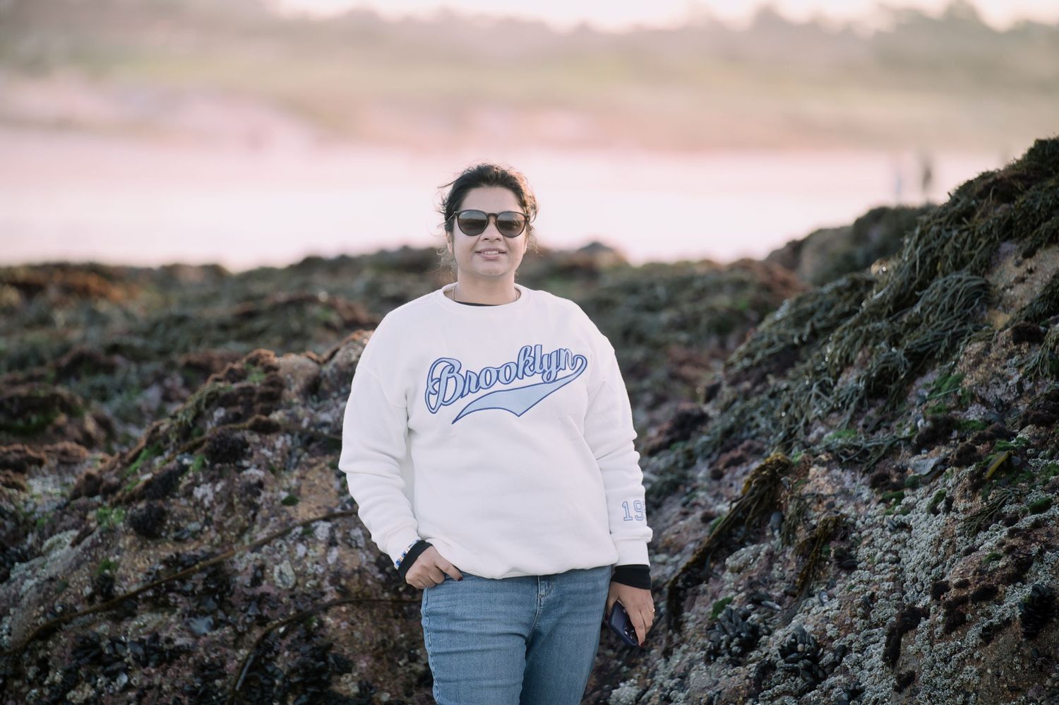 A woman in a white Brooklyn sweatshirt and sunglasses stands on rocky coastal terrain at sunset.
