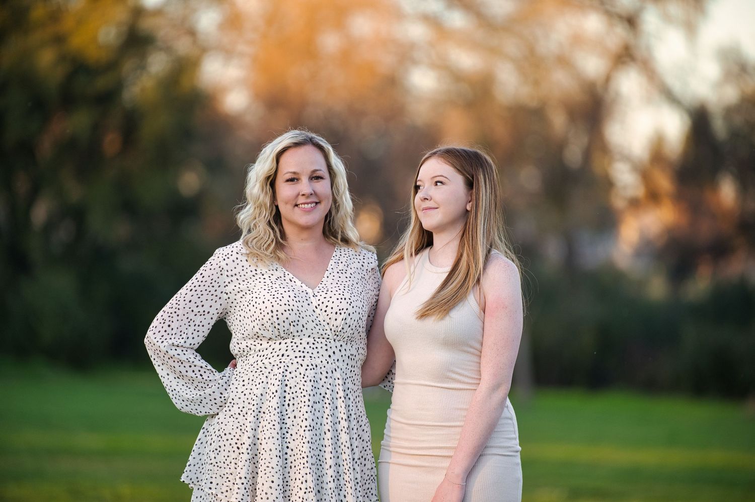 Two blonde women pose together outdoors in park with autumn foliage background.