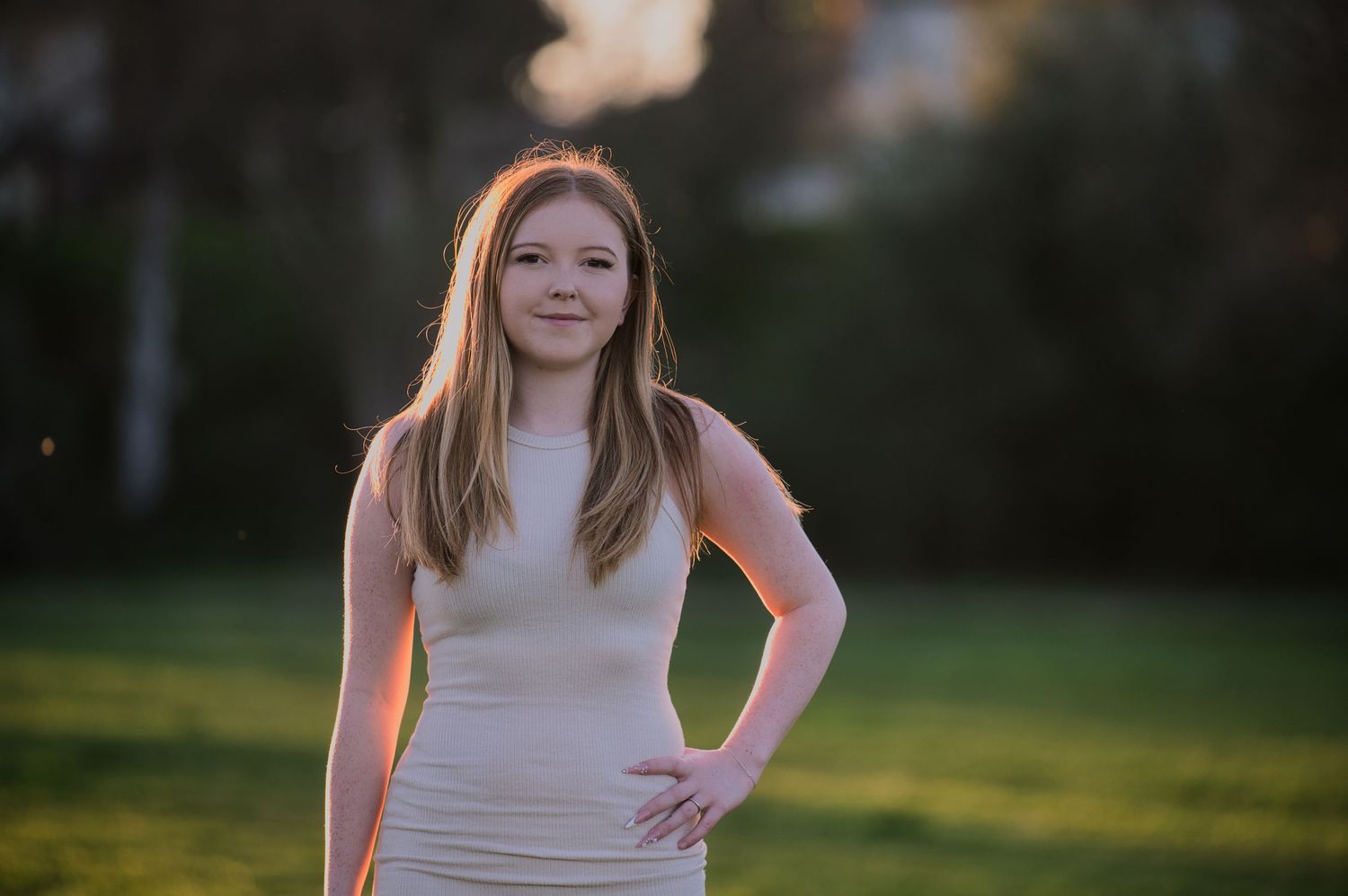 A young teenage girl in a cream dress poses confidently on a green lawn in golden sunlight.