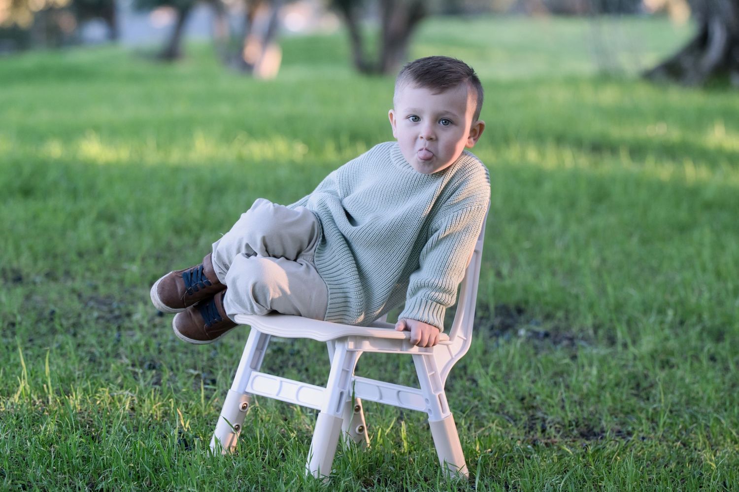 Adorable young boy in sage green knit sweater sitting relaxed on small white wooden chair in sunny green park.