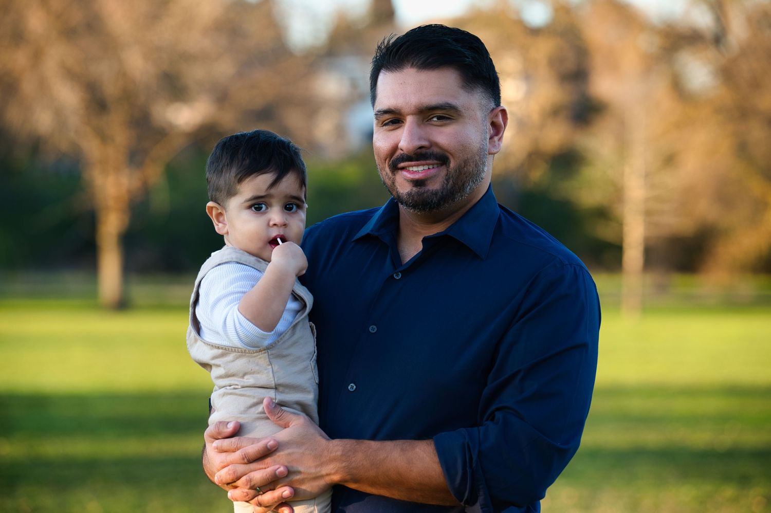 Bearded father in navy shirt holding baby boy outdoors in sunlit park setting.