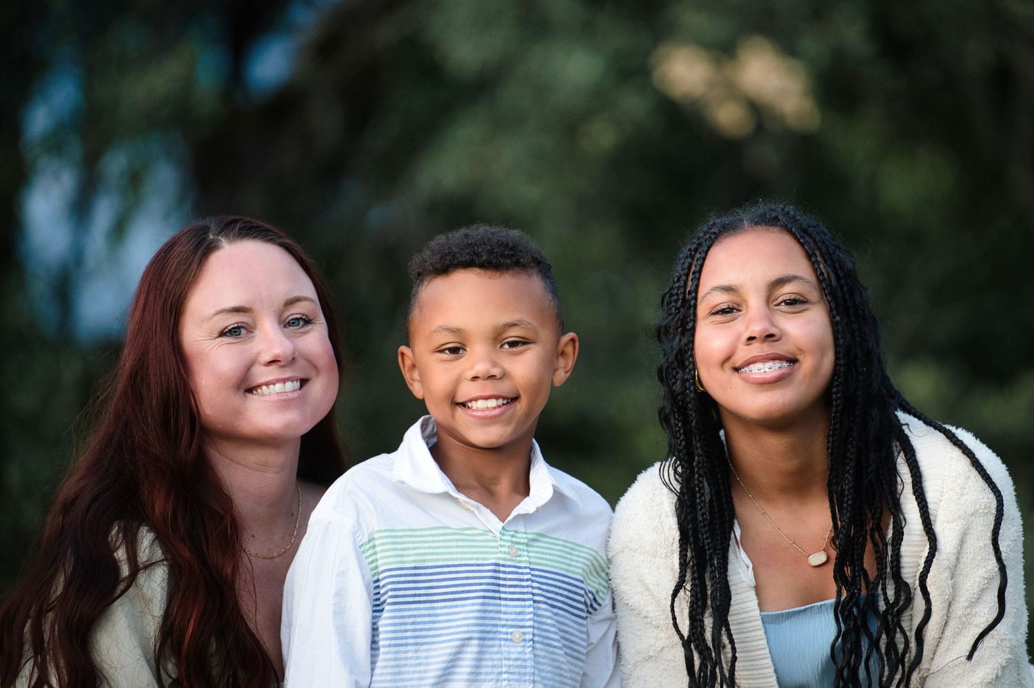A woman with red hair and a teenage girl smile with a young boy between them in an outdoor setting.