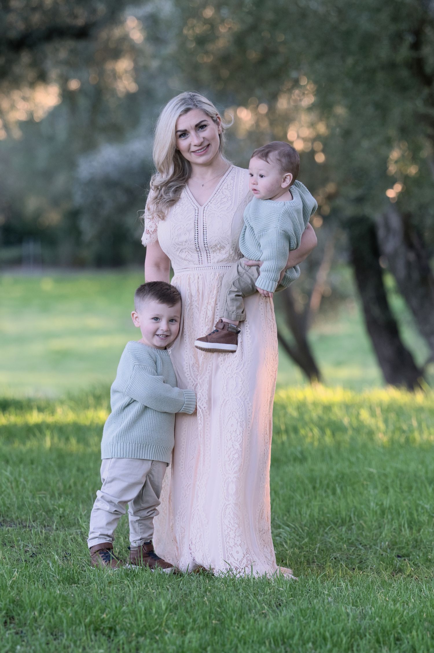 A blonde mother in a pink lace gown holds a baby while a young boy hugs her in a green park.