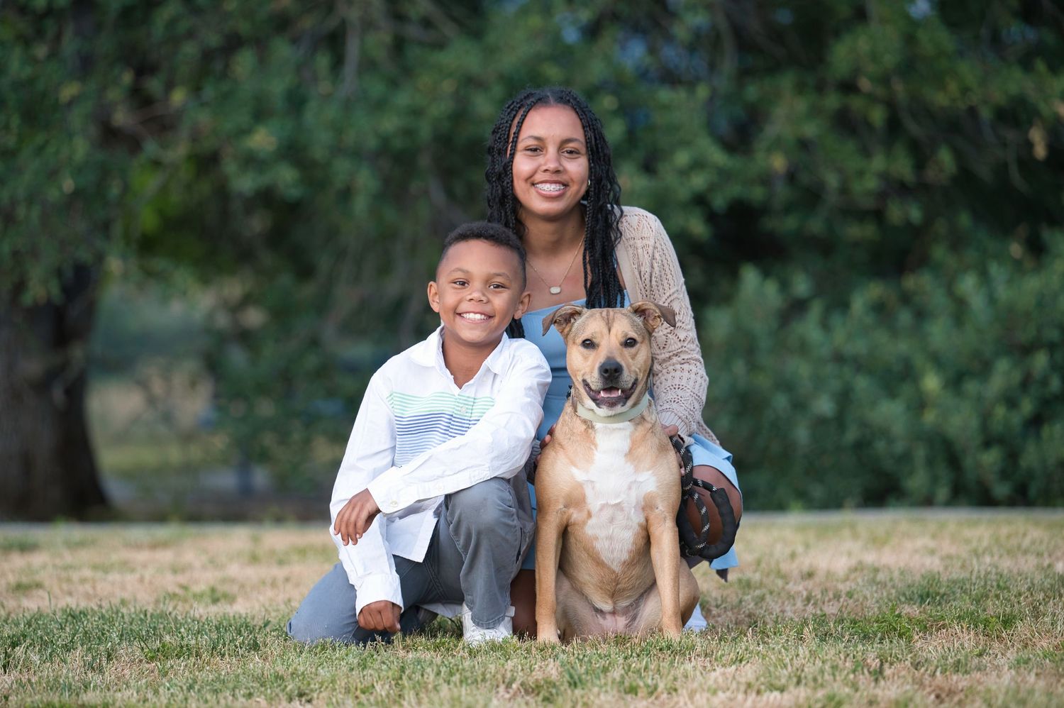 Woman and young boy posing with a tan dog on green grass with trees in the background.
