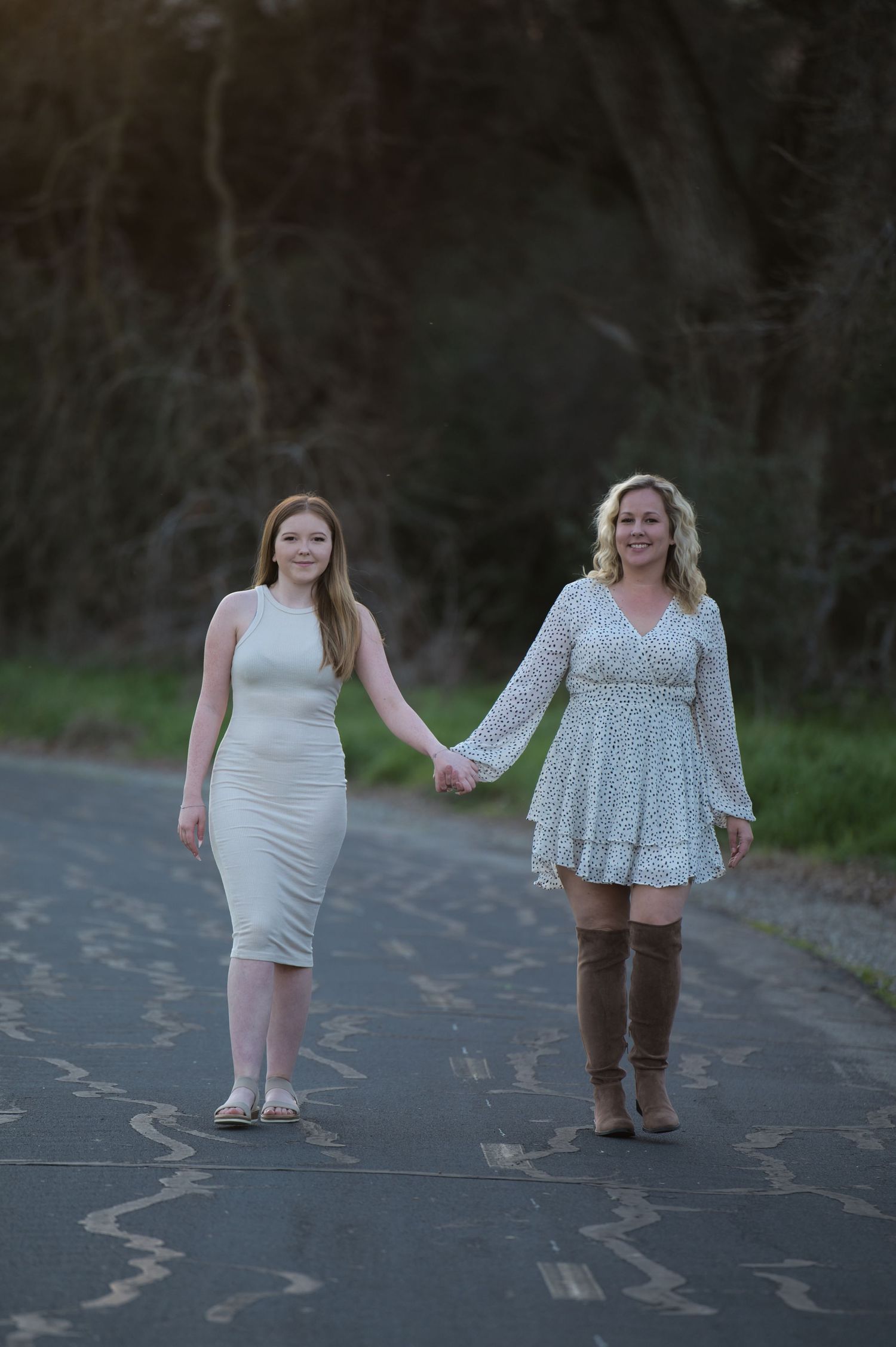 Two women in white dresses hold hands while walking down a quiet road surrounded by trees.