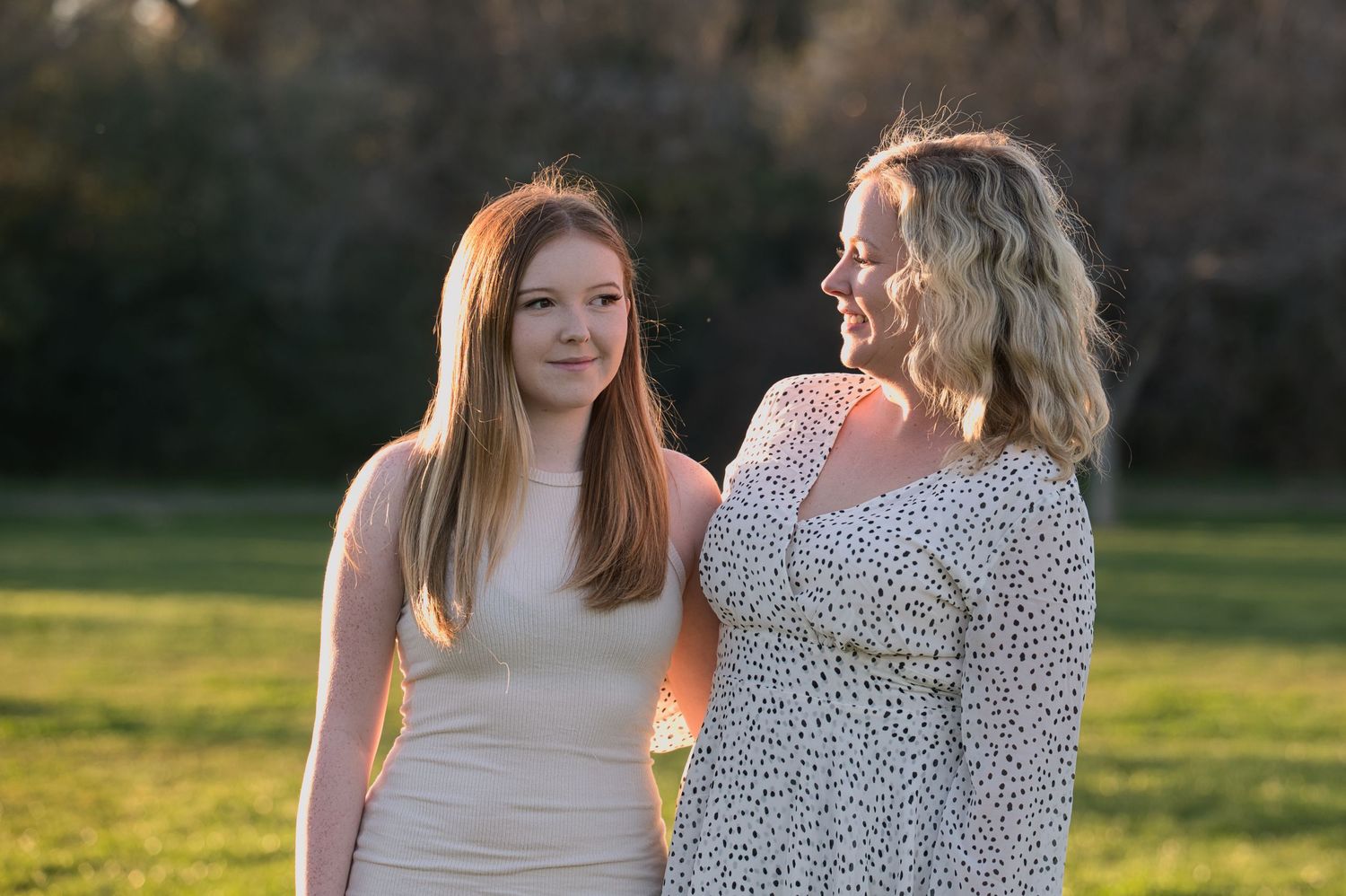 Two women smile at each other warmly in a sunlit outdoor park setting.