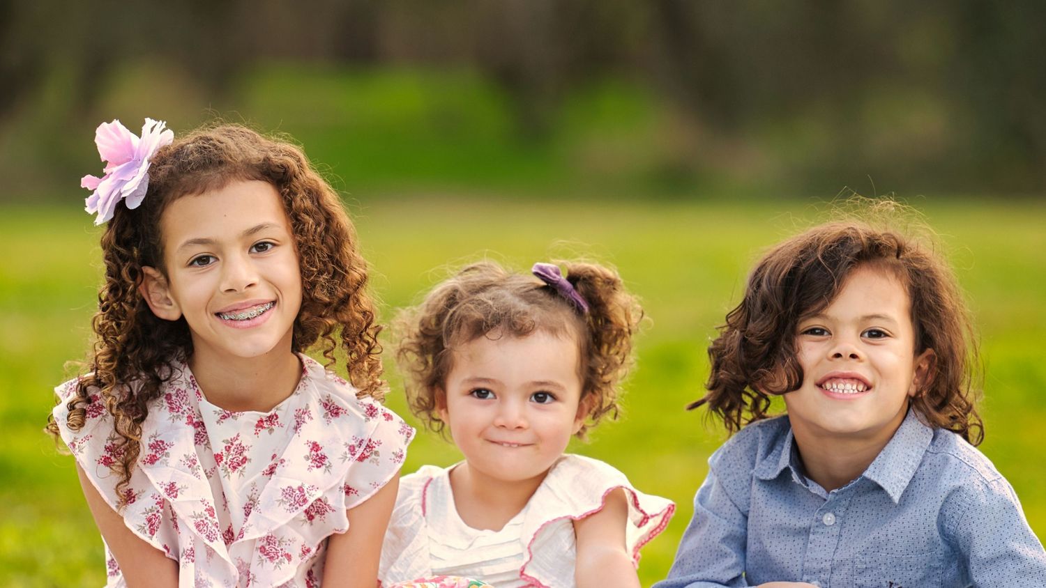 Three young curly-haired siblings sitting together smiling outdoors on bright green grass in spring.