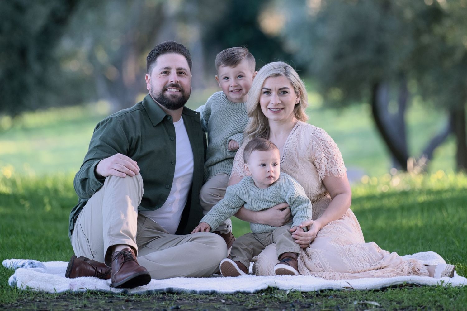 Family of four sitting on white blanket in sunny green park, parents with two young boys in coordinating outfits.