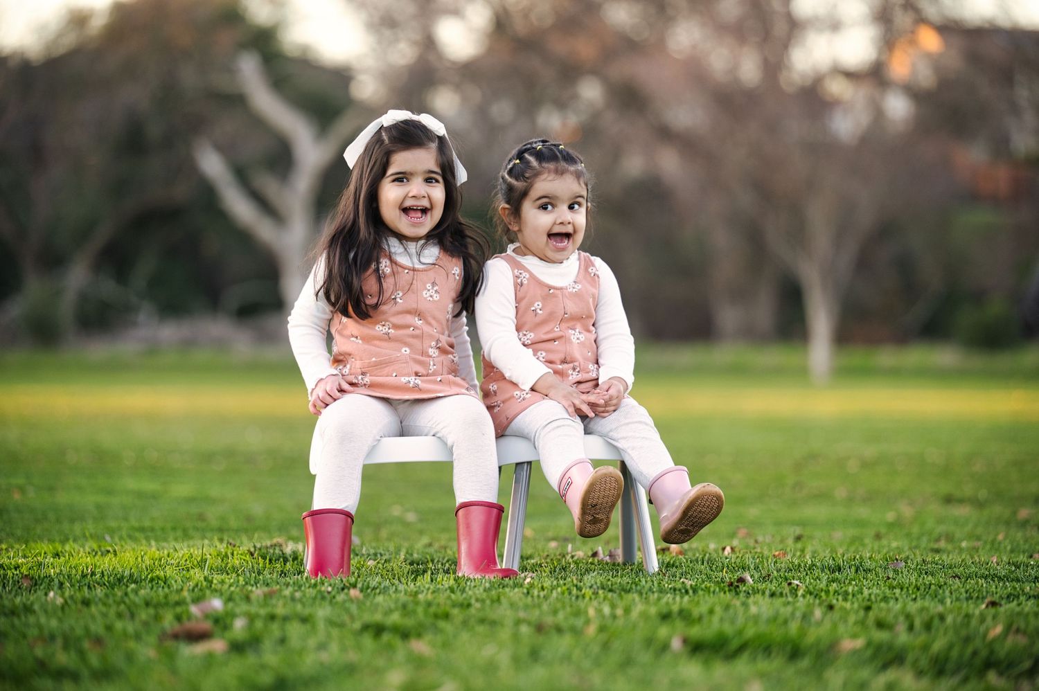 Two young sisters in matching pink dresses laughing while sitting on white chairs in park.