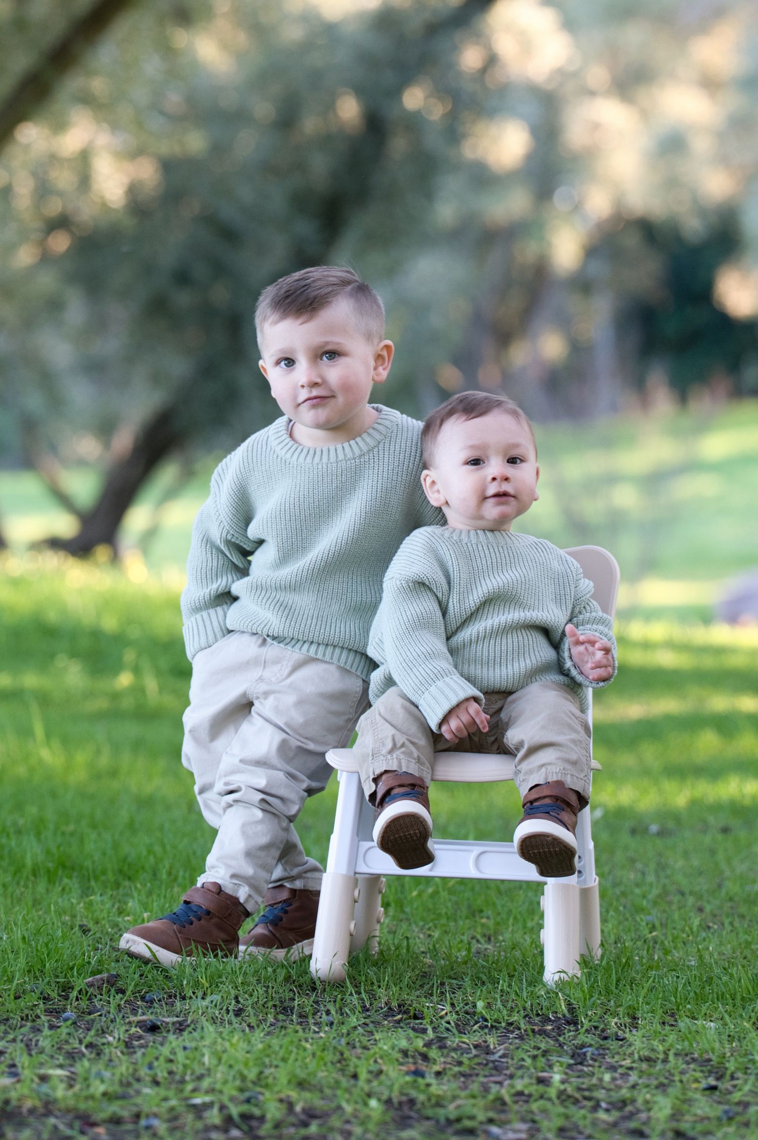 Two young brothers in matching sage green sweaters pose outdoors on a small white chair in a lush green park.