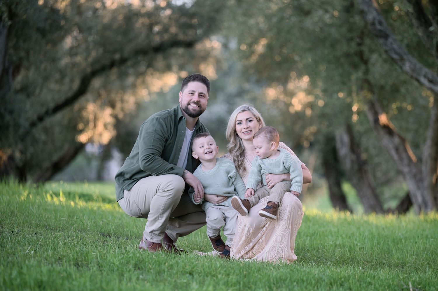 Happy family of four posing outdoors with two young children in matching sage green outfits among olive trees.
