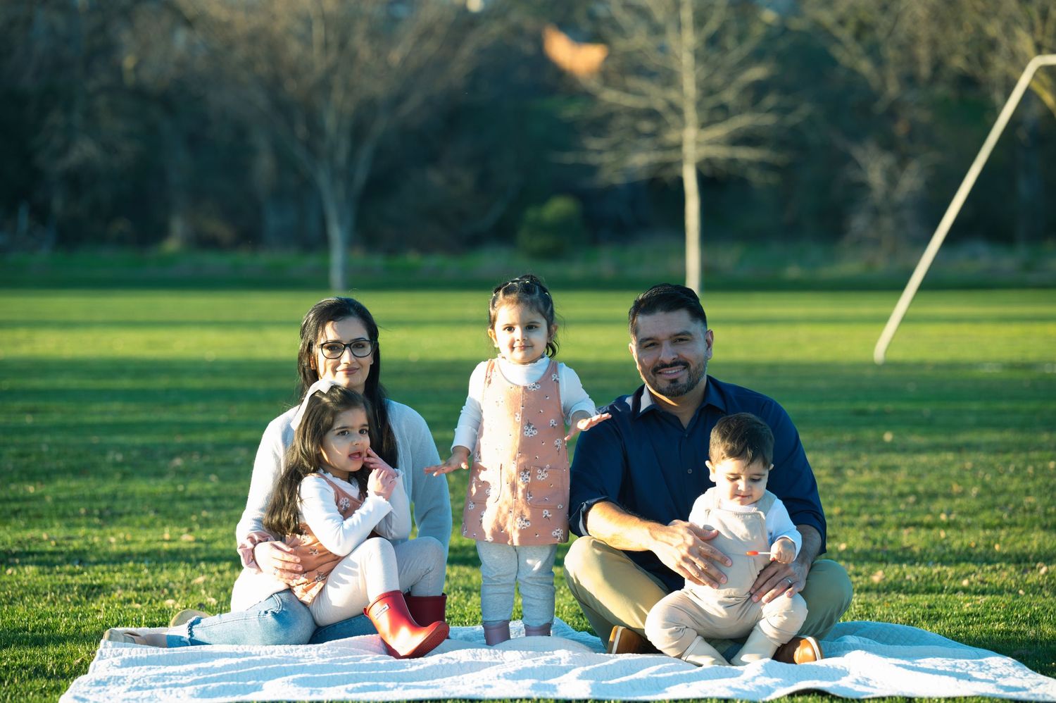 A cheerful family of five enjoys a sunny day sitting on a blanket in a lush green park.
