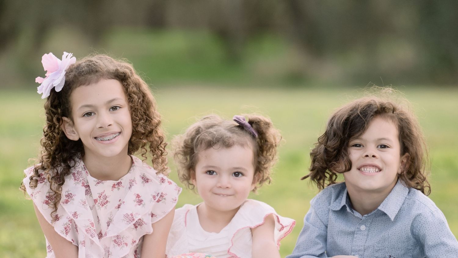 Three young curly-haired siblings smile together on a green lawn in pastel outfits.