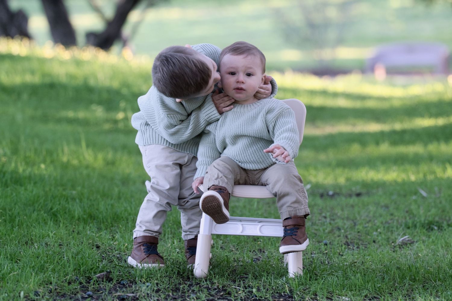 Two young boys in matching sage sweaters share a sweet moment, one whispering to the other outdoors.